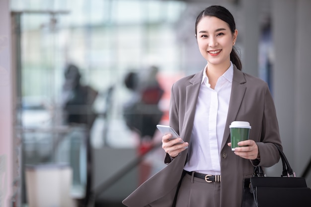 Mujer asiática con smartphone de pie contra la calle edificio borroso. Foto de negocios de moda de hermosa chica en suite casual con teléfono y taza de café