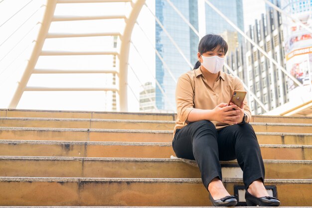 Mujer asiática con sobrepeso con máscara con teléfono y sentado en la ciudad