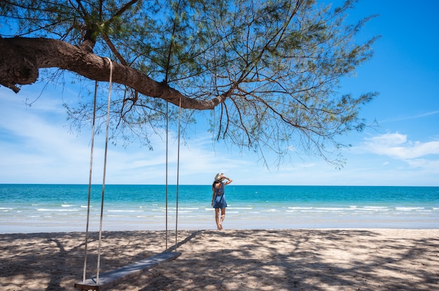 Mujer asiática con sombrero caminando por la playa y columpio de madera colgando de un árbol en el mar tropical