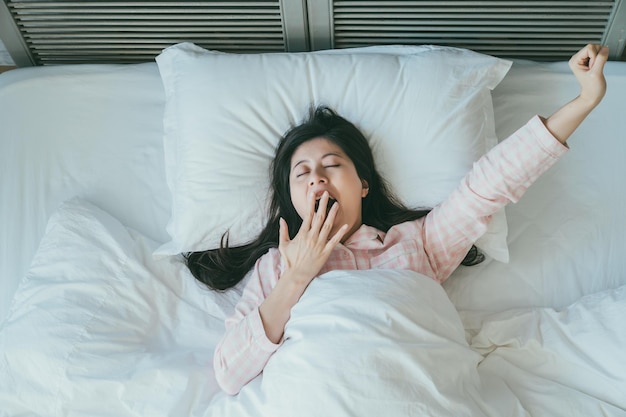 una mujer asiática soñolienta que usa pijama está levantando el brazo y cubriendo su boca mientras bosteza después de despertarse temprano en la mañana en la cama en casa.