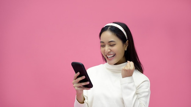 Mujer asiática sonriendo y usando teléfono móvil con tableta y emocionada por comprar en línea en un fondo rosa aislado