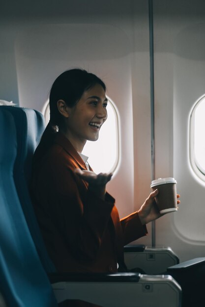 Mujer asiática sonriente disfrutando de un vuelo cómodo con teléfono inteligente mientras está sentada en la cabina del avión Pasajeros cerca de la ventana