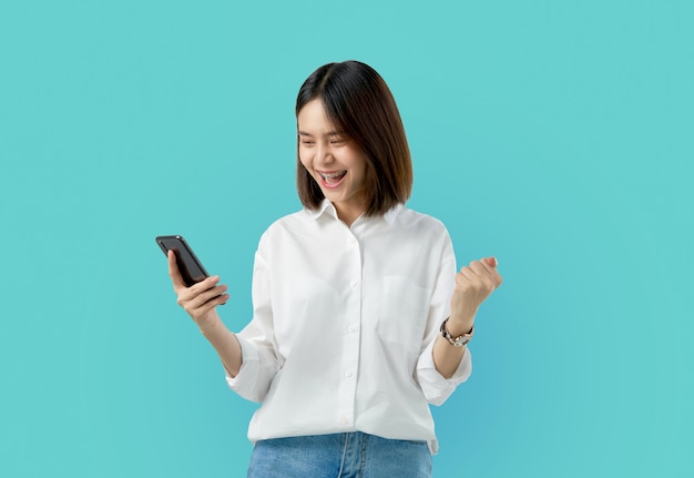 Mujer asiática sonriente de los jóvenes que sostiene el teléfono elegante con la mano del puño y emocionada para el éxito en azul claro