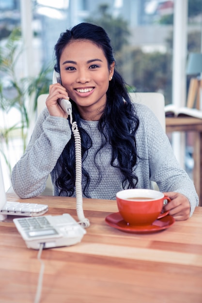 Mujer asiática sonriente en la llamada de teléfono que sostiene la taza en oficina