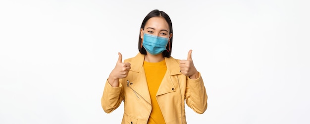 Mujer asiática sonriente con mascarilla médica que muestra el apoyo de los pulgares hacia arriba con equipo de protección de