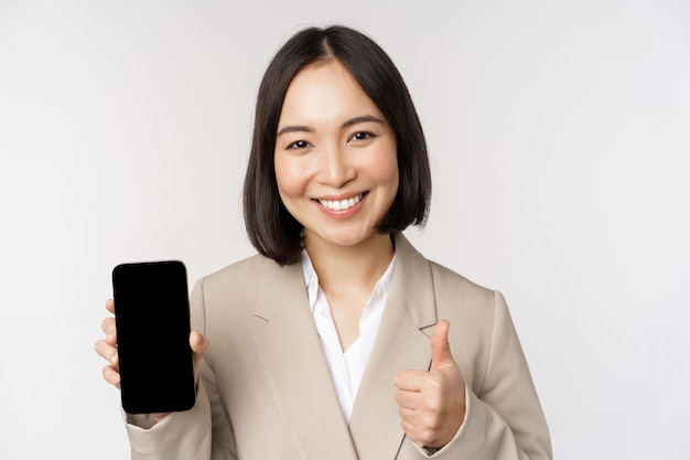 Mujer asiática sonriente que muestra la pantalla del teléfono inteligente y los pulgares hacia arriba La persona corporativa demuestra la interfaz de la aplicación del teléfono móvil de pie sobre fondo blanco