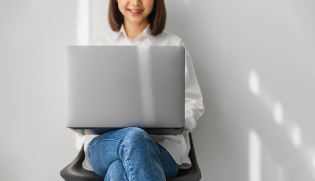 Mujer asiática sonriente que se sienta en silla y que usa la computadora portátil en casa.