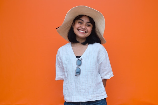 Mujer asiática sonriente con ropa informal de verano, sombrero y gafas de sol mirando la cámara en el estudio naranja b