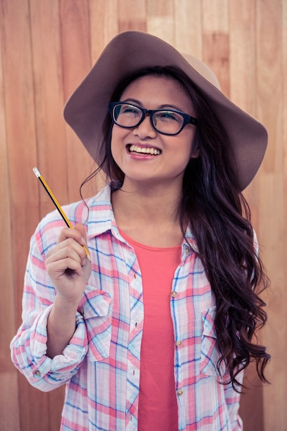 Mujer asiática sonriente con el sombrero que sostiene el lápiz contra la pared de madera