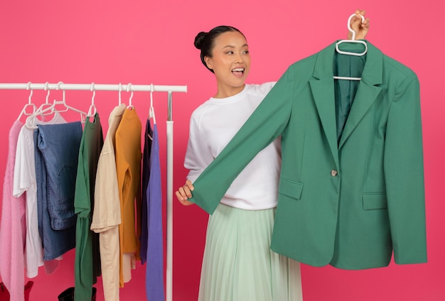 Mujer asiática sonriente sosteniendo un blazer verde mientras está de pie cerca del estante de ropa