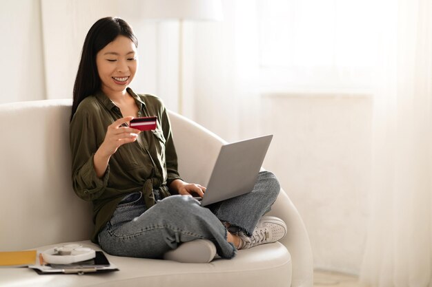 Mujer asiática sonriente usando laptop y tarjeta bancaria en casa