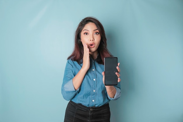 Mujer asiática sorprendida con camisa azul apuntando a su teléfono inteligente aislado por fondo azul