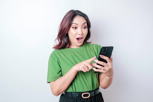 Mujer asiática sorprendida con camiseta verde apuntando a su teléfono inteligente aislado de fondo blanco