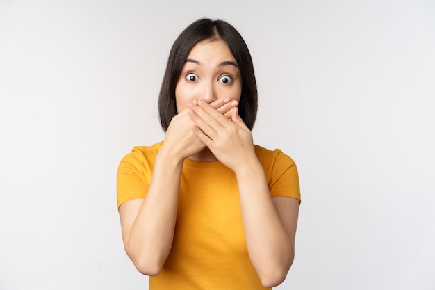 Una mujer asiática sorprendida cubre la boca con las manos mirando sorprendida con una expresión facial sin palabras de pie en una camiseta amarilla contra el fondo blanco