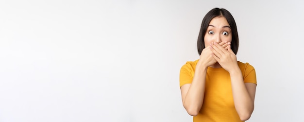 Una mujer asiática sorprendida cubre la boca con las manos mirando sorprendida con una expresión facial sin palabras de pie en una camiseta amarilla contra el fondo blanco