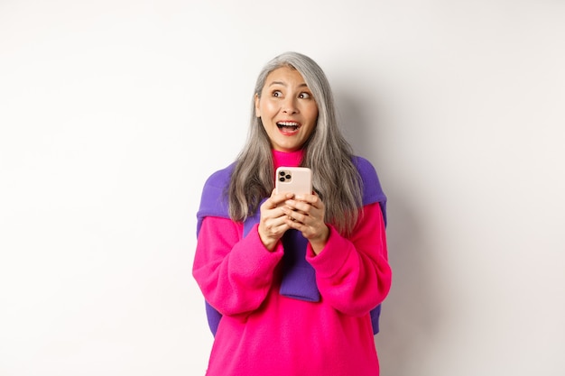 Foto mujer asiática sorprendida sonriendo y mirando a un lado fascinado después de leer la promoción en el teléfono inteligente, de pie con el teléfono móvil sobre fondo blanco.