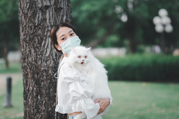 Mujer asiática sosteniendo un gato blanco en un parque con máscaras antivirus durante el brote de coronavirus