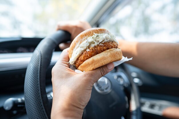 Mujer asiática sosteniendo una hamburguesa para comer en un auto peligroso y arriesgarse a un accidente