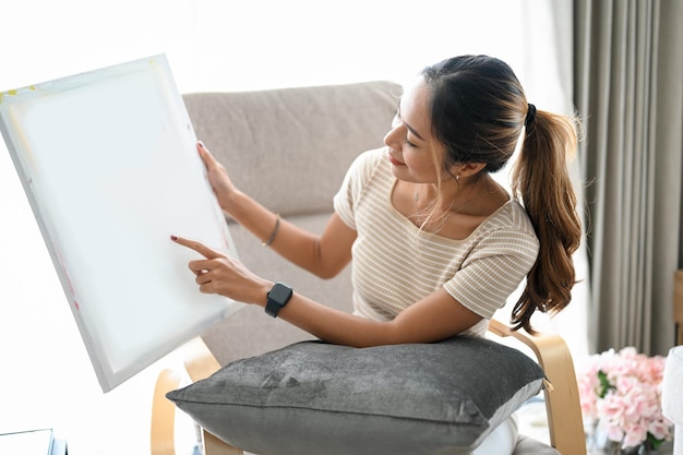 Una mujer asiática sosteniendo un lienzo en blanco mirando un lienzo o mostrando su imagen en un lienzo