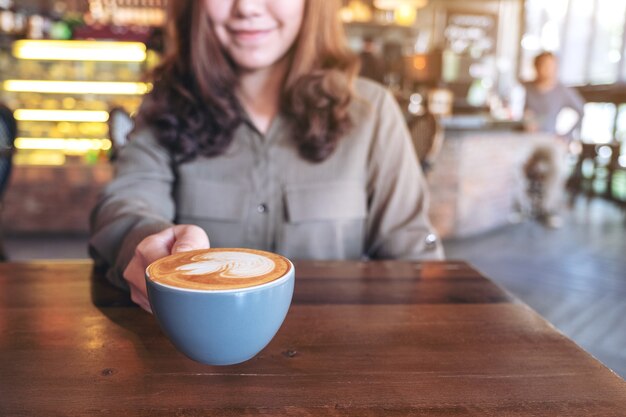 Mujer asiática sosteniendo una taza azul de café con leche caliente con arte latte en la mesa de madera en el café