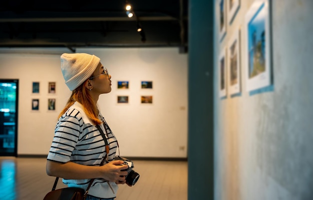Foto mujer asiática sostiene la cámara en la colección de la galería de arte frente a cuadros enmarcados