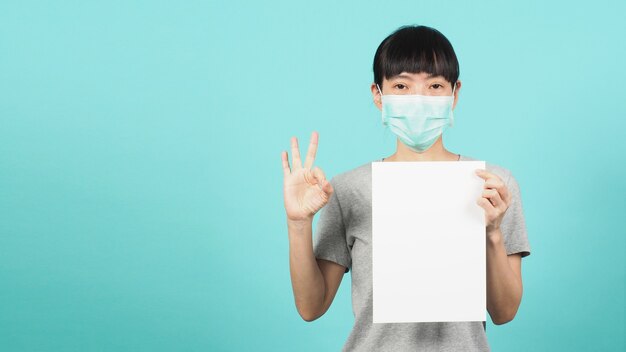 La mujer asiática sostiene papel A4 en blanco y hace bien la señal de mano y usa mascarilla o máscara médica sobre fondo azul y verde o menta.