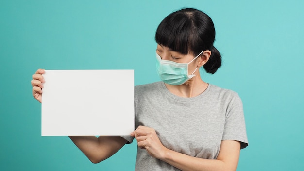 La mujer asiática sostiene papel A4 en blanco y usa mascarilla o máscara médica sobre fondo azul y verde o menta.