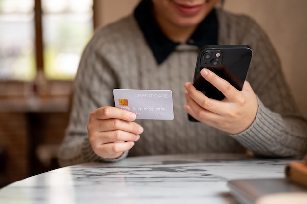 Una mujer asiática con su teléfono inteligente y una tarjeta de crédito sentada en una mesa en el interior