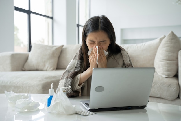 Una mujer asiática se suena la nariz en una servilleta se sienta en un escritorio de oficina en casa estudia en una computadora portátil un enfermo