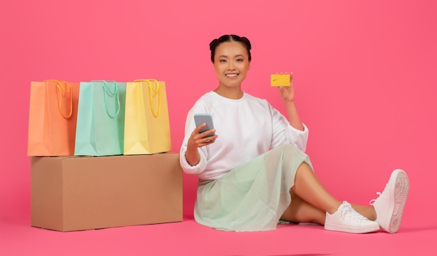 Mujer asiática con teléfono inteligente y tarjeta de crédito sentada cerca de bolsas de compras