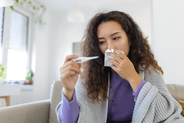 Una mujer asiática tiene gripe y está usando un termómetro Enferma con rinitis Una mujer con hiyab tiene goteo nasal Mujer enferma con gripe acostada en el sofá mirando la temperatura en el termómetro