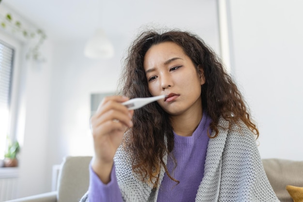 Una mujer asiática tiene gripe y está usando un termómetro enferma con rinitis una mujer con hiyab tiene goteo nasal mujer enferma con gripe acostada en el sofá mirando la temperatura en el termómetro