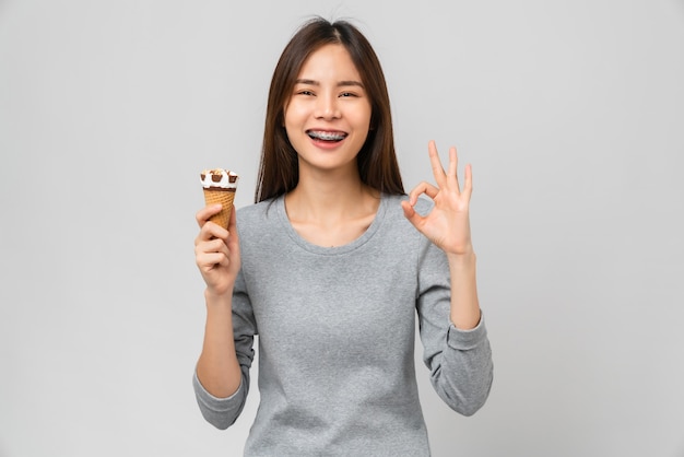 Mujer asiática con tirantes sosteniendo helado y muestra signo de ok sobre fondo gris.