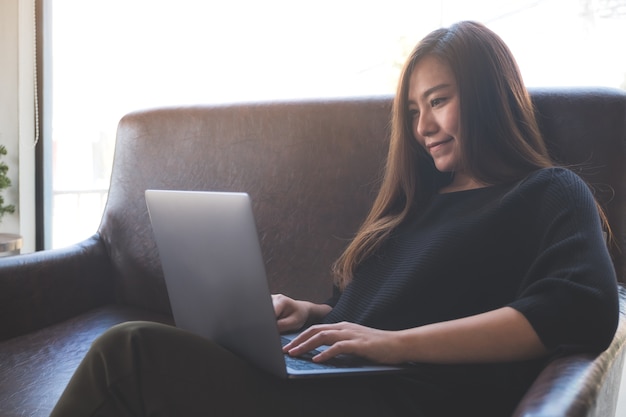 una mujer asiática trabajando y escribiendo en el teclado del ordenador portátil mientras está sentado en el sofá