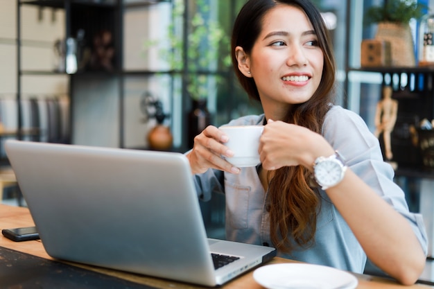 Mujer asiática trabajando y tomando café en la cafetería con computadora portátil sonrisa y trabajo feliz