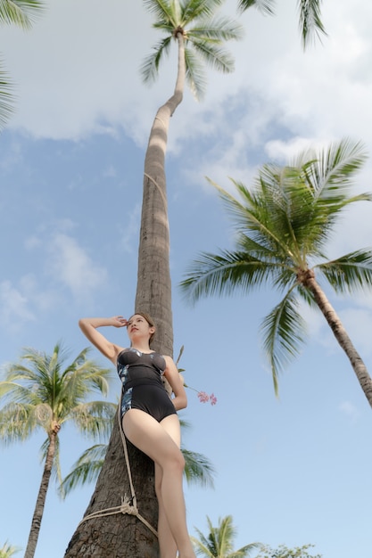 Mujer asiática en traje de baño de una pieza que publica con palmera de coco en playa tropical