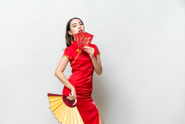 Mujer asiática con traje de cheongsam oriental sosteniendo sobres rojos Ang Pow y fan posando en un estudio gris claro para conceptos de año nuevo chino, textos extranjeros significan gran suerte gran ganancia