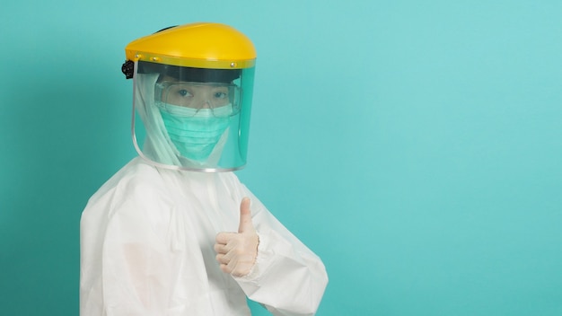 Foto mujer asiática en traje de ppe, cara encogida, guantes médicos y mascarilla con el pulgar hacia arriba o como signo de mano en verde, fondo azul tiffany.