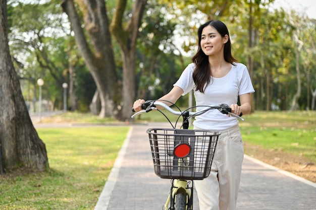 Mujer asiática tranquila caminando por la acera con su bicicleta en el hermoso parque natural