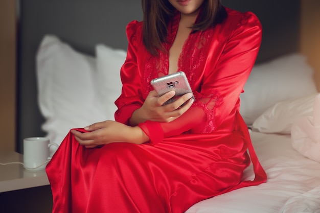 Mujer asiática en túnicas de satén rojo con un teléfono móvil mientras está sentado en una cama por la noche