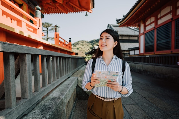 mujer asiática turista sosteniendo un mapa de papel caminando haciendo turismo en el templo kiyomizu dera kyoto japón en vacaciones de verano. joven viajera sonriendo visitando un santuario japonés de madera roja en el pasillo.