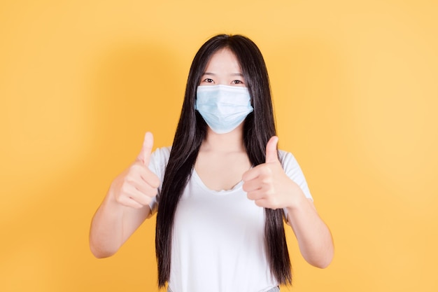 Foto mujer asiática usa camiseta blanca con mascarilla médica para proteger el coronavirus covid19 mostrando dos pulgares dobles hacia arriba sobre fondo amarillo