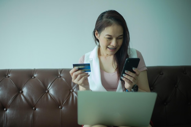 Mujer asiática usa laptop y teléfono inteligente con tarjeta de crédito en el sofá