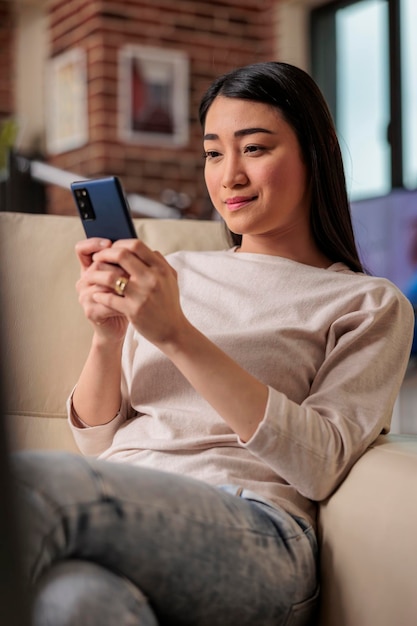 Mujer asiática usuaria de smartphone en casa en el sofá, mirando el teléfono con una sonrisa auténtica. Usuario móvil de tecnología web de Internet