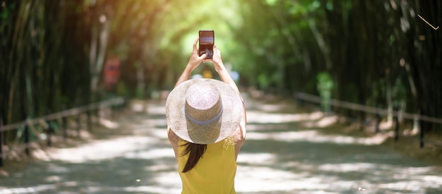 Mujer asiática en vestido amarillo y sombrero Viajando en el túnel de bambú verde Viajero feliz tomando fotos por teléfono móvil en el templo Chulabhorn wanaram en Nakhon Nayok Tailandia