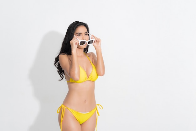 Mujer asiática con un vestido de bikini amarillo de manera permanente, con gafas de sol blancas, en una moda de verano en el estudio de disparo fondo blanco.