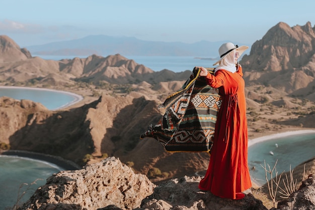 Mujer asiática en vestido y sombrero de verano de pie en la cima de la colina y sosteniendo kain songket en Padar Island L