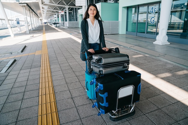 Mujer asiática viajera rodando carrito de equipaje fuera del aeropuerto de osaka con una sonrisa segura en la cara. señorita con carro de equipaje caminando afuera en verano relajante esperando transporte de vuelo.