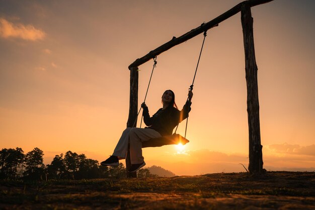 Mujer asiática viajera de silueta sentada con un columpio de madera en la cima de la montaña al atardecer