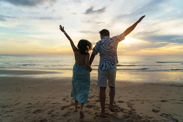 Mujer asiática viajera sosteniendo la mano del hombre y mirando hermosa puesta de sol en la playa Pareja de vacaciones en verano y concepto de libertad